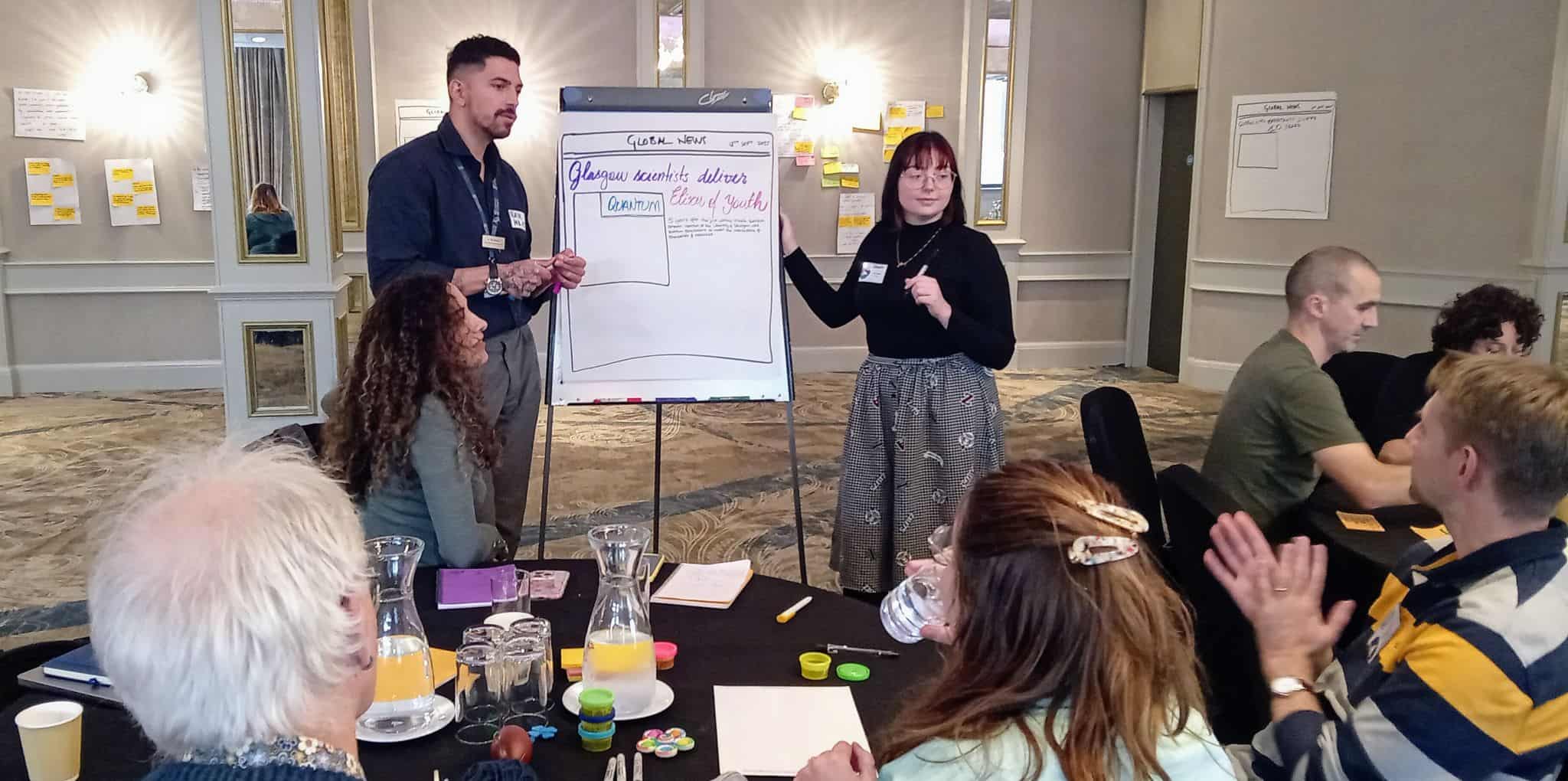 Amy Eden (DiveIn PGR - cohort 1) and Rair Macedo (DiveIn supervisor) present a project idea standing beside a flipchart with a fake news paper headline at the Project Scope and Connect Workshop 2025.