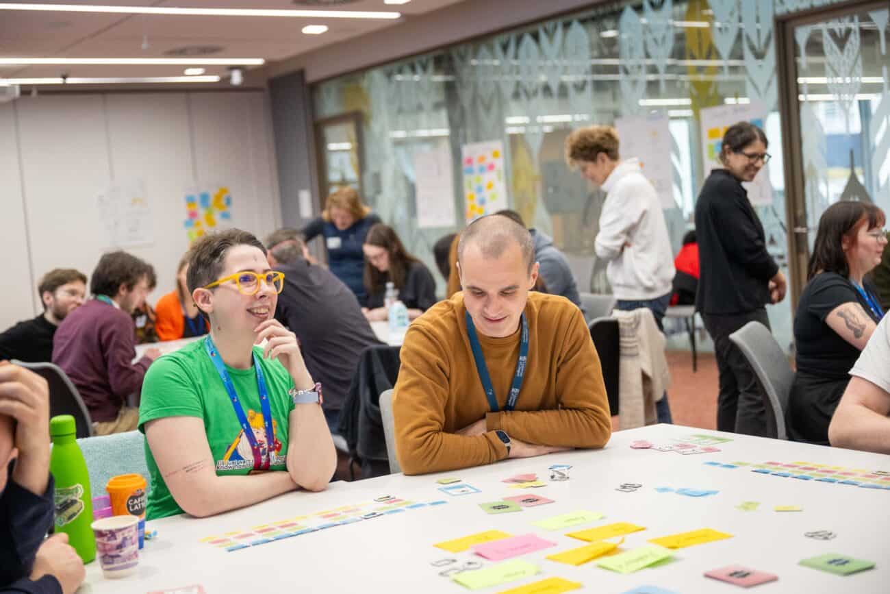 A group of people sit around a table in a bright, modern room, working with colourful cards, post‑it notes, and printed materials as part of a collaborative workshop activity. Additional groups are gathered at tables in the background, and large windows and whiteboards with notes and diagrams line the walls.