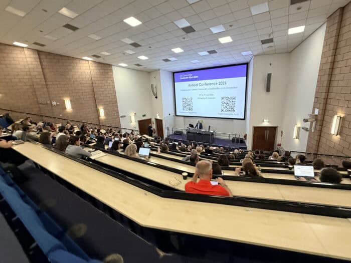 Lecture theatre packed with conference delegates viewing a large screen displaying "UK Council for Graduate Education, Annual Conference 2026, Postgraduate Cultures: Community, Collaboration and Connection, 2nd & 3rd July 2026, University of Liverpool" with two QR codes for the conference programme and digital delegate pack.