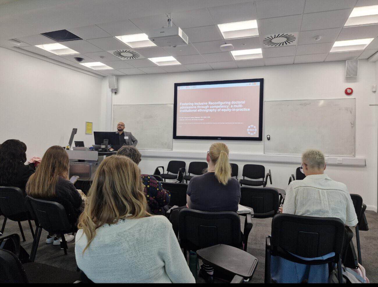 Qammer H. Abbasi presenting to a small group of seated attendees, with a screen behind him titled "Fostering Inclusive Reconfiguring doctoral admissions through competency: a multi-institutional ethnography of equity-in-practice."