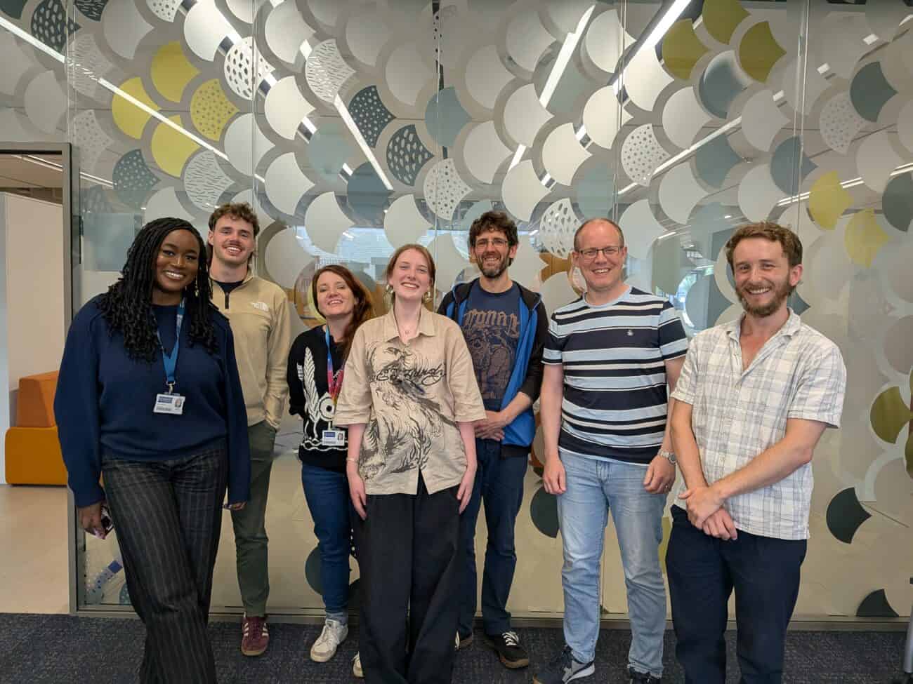 Group photo of In2research summer placement participants, DiveIn CDT placement hosts and directors standing together in a modern university building, in front of a large glass wall with an abstract pattern. Several people are wearing lanyards and everyone is facing the camera.